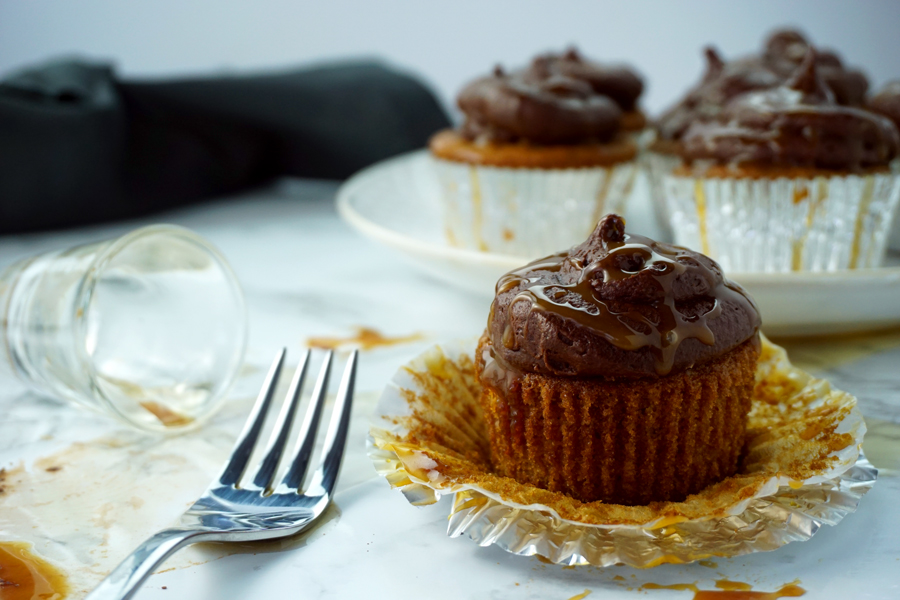 A caramel-flavored cupcake with chocolate buttercream and caramel drizzled on top sits in the foreground of a bottle of Guinness and a shot glass of Jameson Irish Whiskey