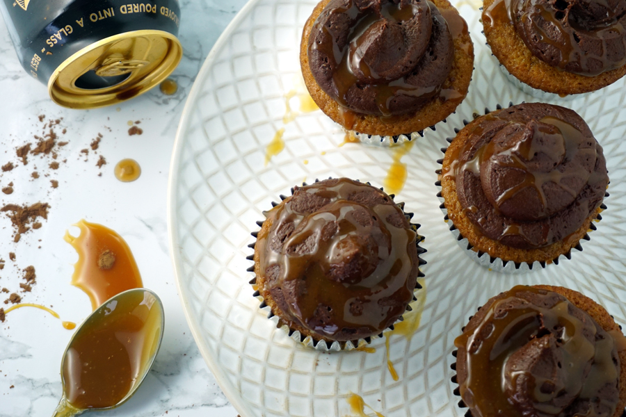 A caramel-flavored cupcake with chocolate buttercream and caramel drizzled on top sits in the foreground of a bottle of Guinness and a shot glass of Jameson Irish Whiskey