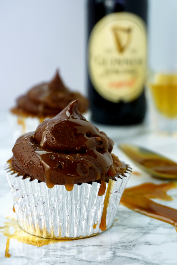 A caramel-flavored cupcake with chocolate buttercream and caramel drizzled on top sits in the foreground of a bottle of Guinness and a shot glass of Jameson Irish Whiskey