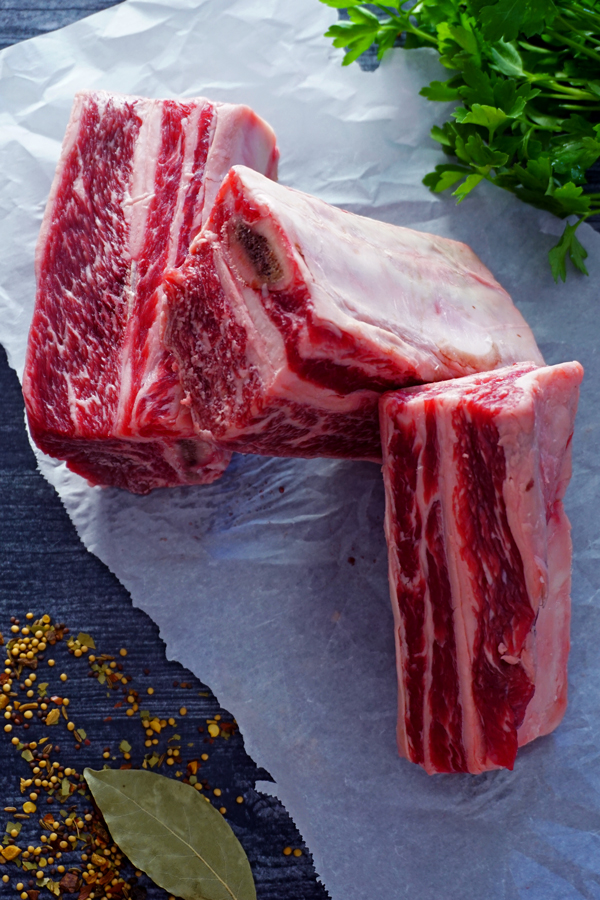 three english-style beef short ribs on a piece of parchment paper with bay leaves, parsley and pickling spices