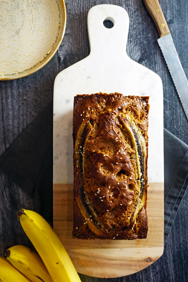 loaf of banana bread on a wood and marble board with a plate, knife and bananas around it