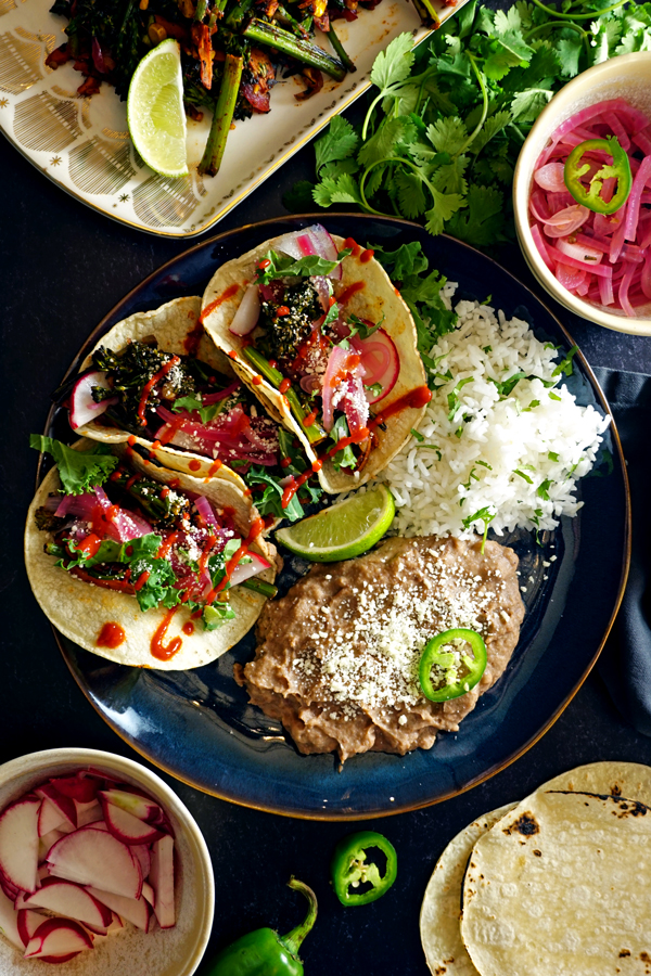 a blue plate with three charred vegetable tacos, rice and beans surrounded by tortillas and other tacos fillings and garnishes