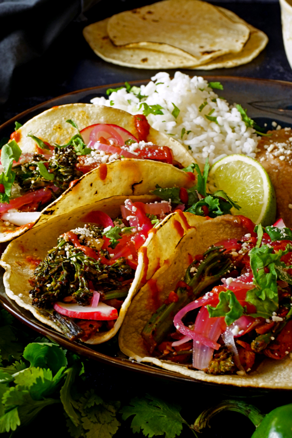 charred vegetables tacos up close on a plate surrounded by cilantro with rice and tortillas in the background