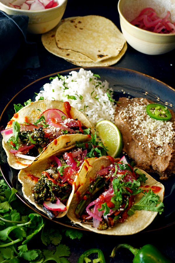 three charred vegetable tacos on a plate with broccolini, radish and cotija cheese and a side of rice and beans