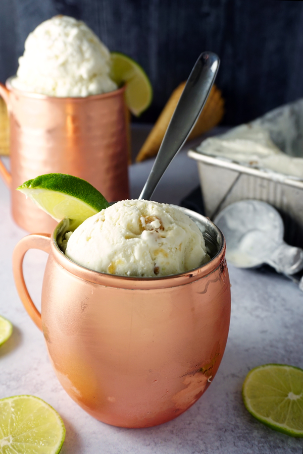 scoops of moscow mule no-churn ice cream in a copper mug, garnished with a lime wedge with another mug and extra ice cream in the background